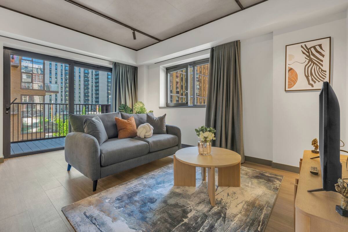 A modern living room with a grey sofa, a round wooden coffee table, and a large window with curtains.