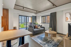 A modern living room with a grey sofa, a round coffee table, and a dining table. There are large windows with curtains, a view of greenery outside, and a piece of abstract art on the wall. - Vibrant Apartments at Luna near Wembley Park by MySquare (Wembley)
