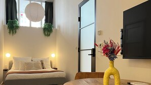 Ein modernes Hotelzimmer mit einem Bett, einem Esstisch mit Stühlen, einem an der Wand montierten Fernseher und einem Fenster mit Vorhängen und Pflanzen.