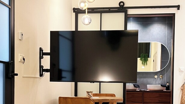 Eine moderne Küche mit einem großen, schwarzen Fernseher an der Wand, einem Holztisch mit zwei Stühlen und einem Holzboden im Fischgrätmuster.
