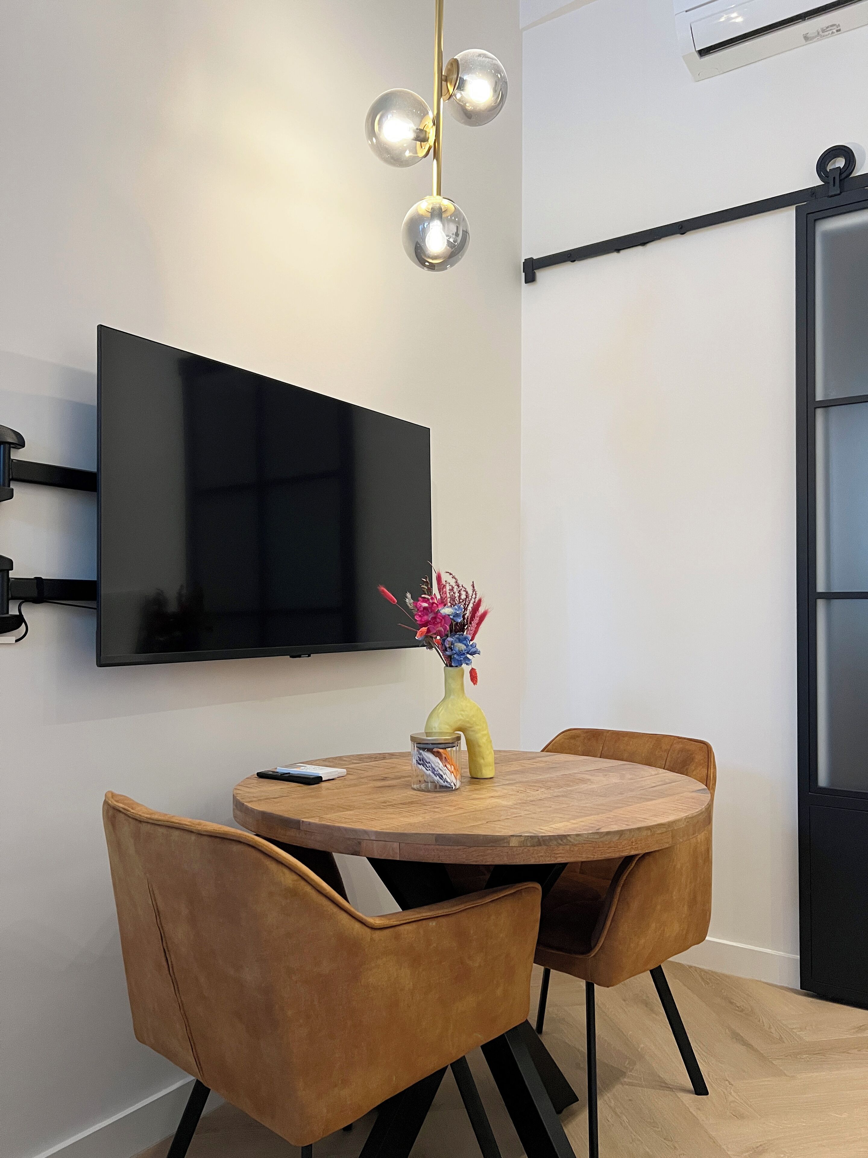 A modern dining area with a round wooden table, two brown chairs, a vase with flowers, and a mounted flat-screen TV.