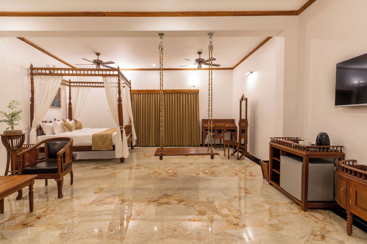 A spacious room with a four-poster bed, a desk, a chair, a small refrigerator, and a television on the wall.
