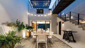 An outdoor patio with a wooden table and chairs, surrounded by potted plants and string lights.