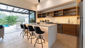 A modern kitchen with a central island, bar stools, wooden shelves, and a view of the outdoors.