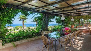 A covered outdoor dining area with a view of the sea, surrounded by lush greenery and potted plants. - Charming One-Bedroom Apartment with Sea View in Villa with Pool by the Sea (Forio)