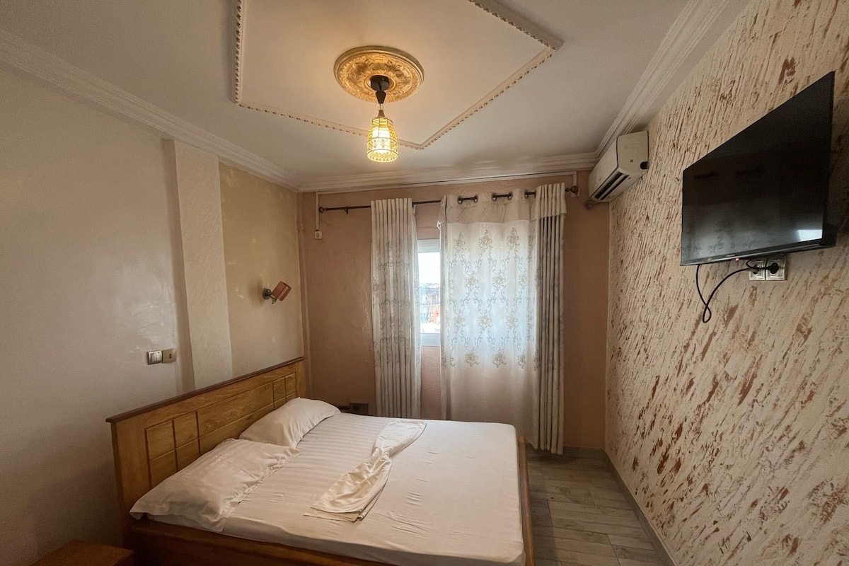 A bedroom with a wooden bed, a ceiling light fixture, a flat-screen TV mounted on the wall, and a window with curtains.
