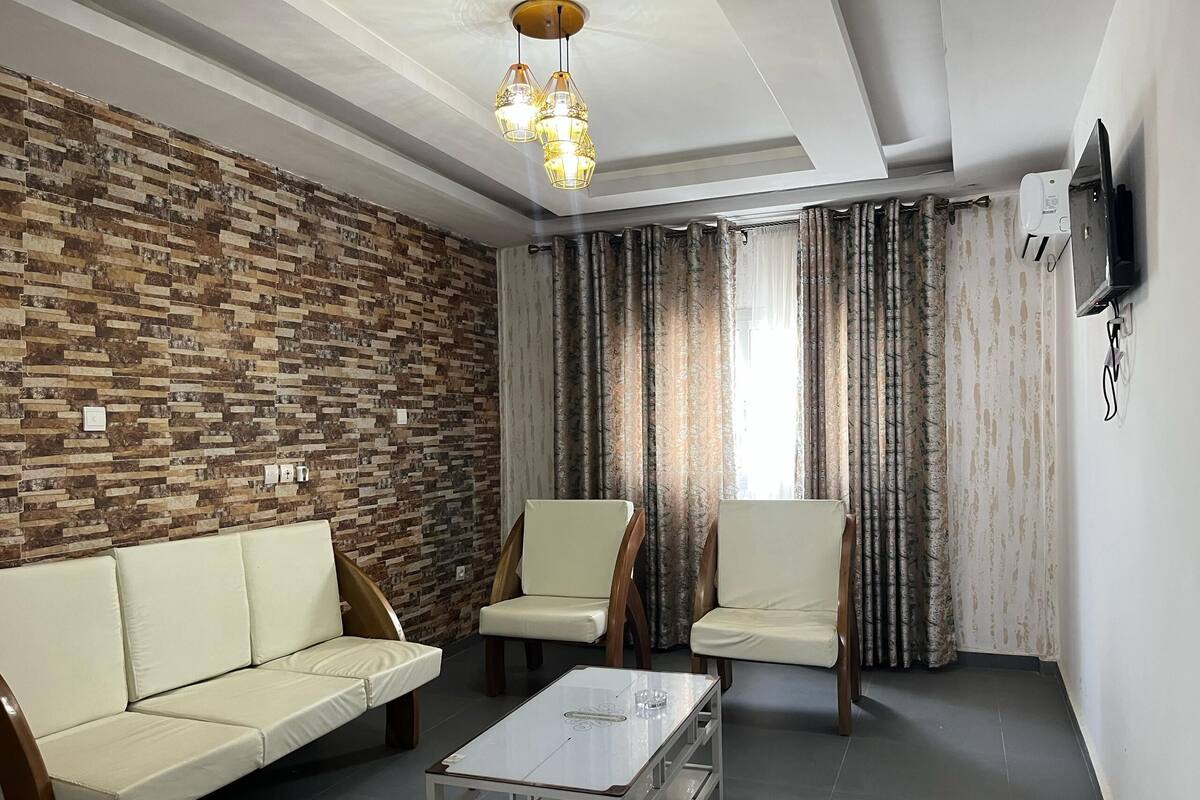 A living room with a brick-patterned wall, a sofa, two chairs, a coffee table, and a TV mounted on the wall.