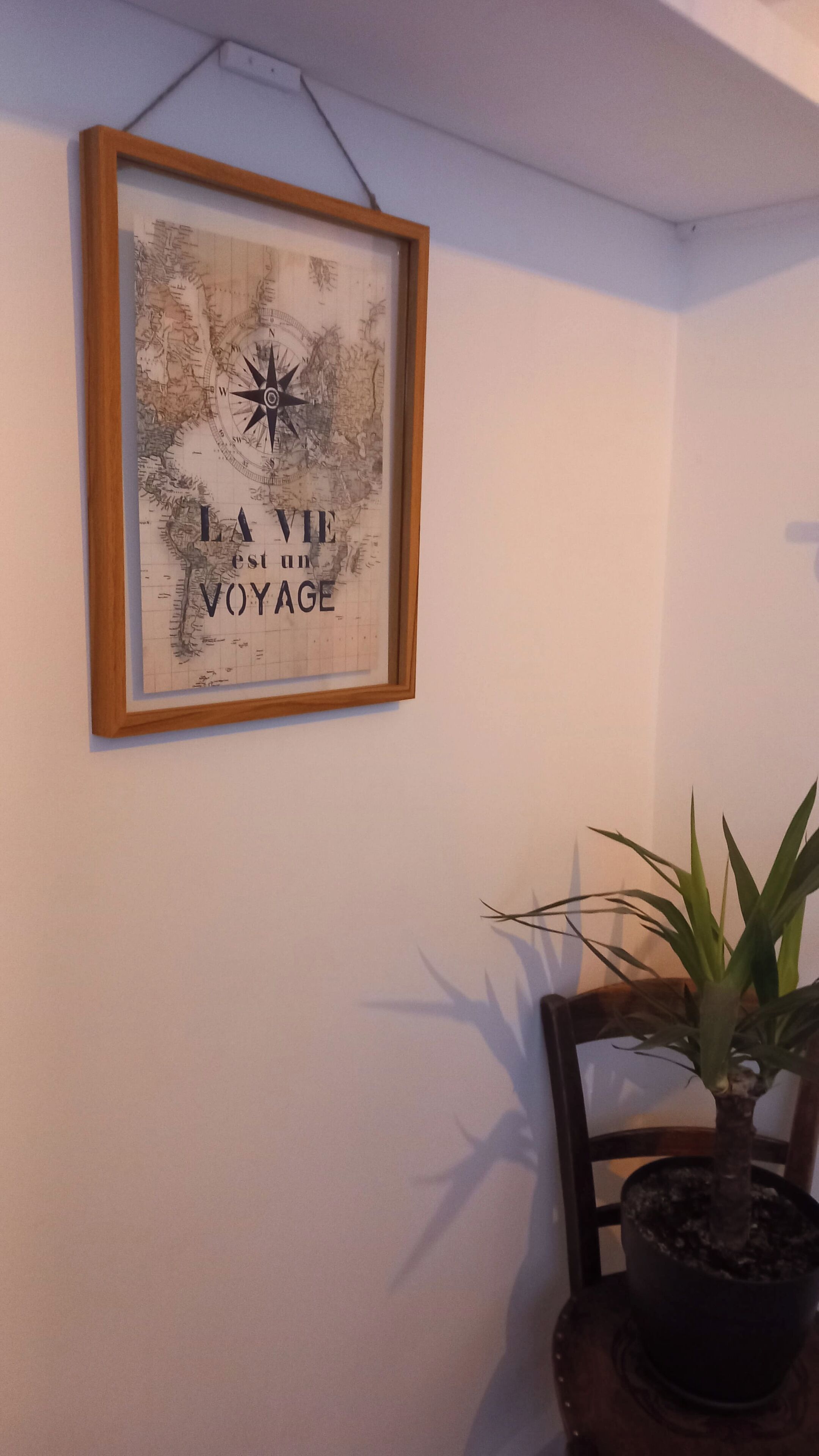 Een lijst met een kunstwerk waarop een kompas en de tekst 'LA VIE EST UN VOYAGE' staan, hangt aan een muur.