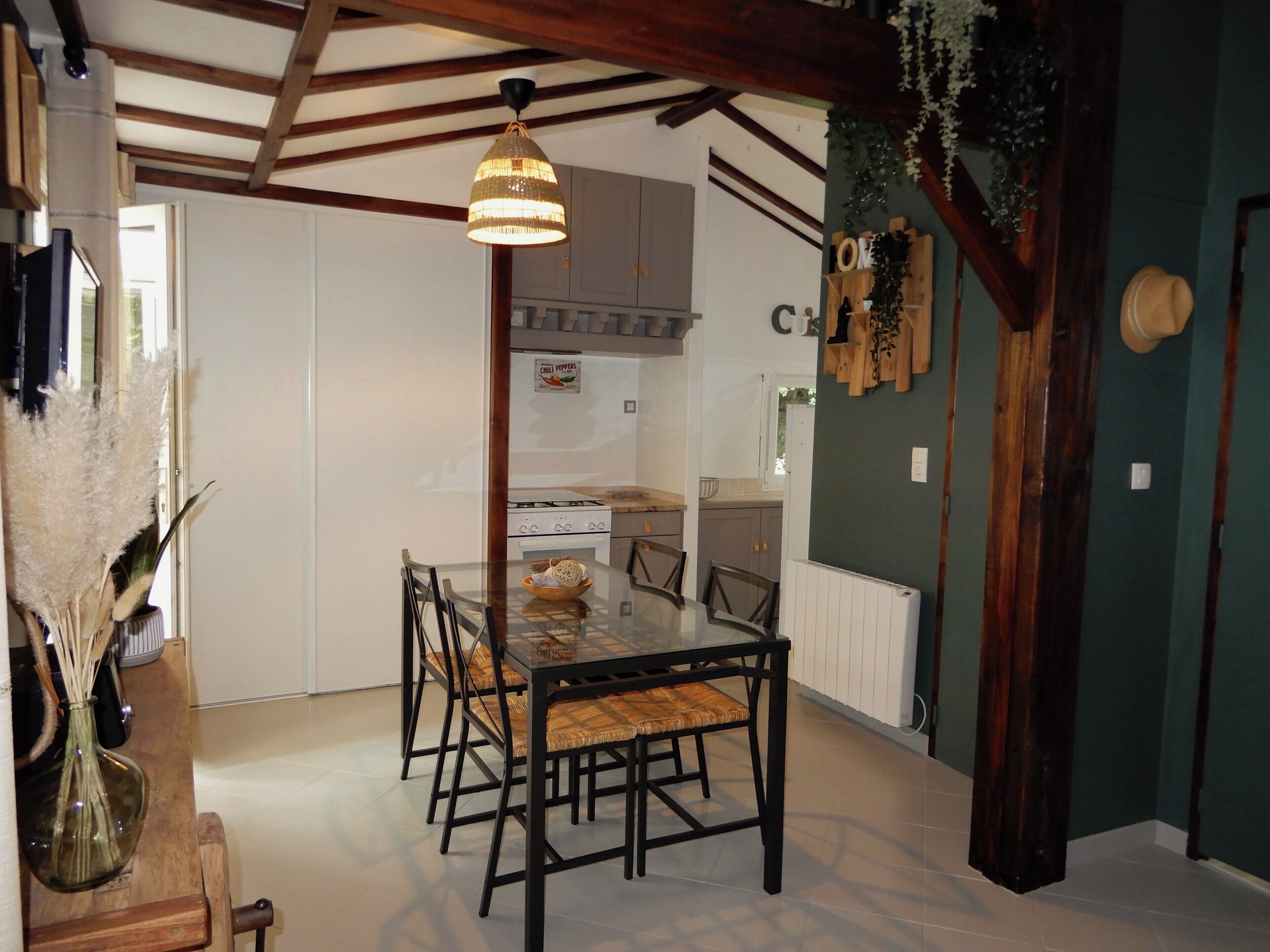 A dining area with a glass table and chairs, a hanging light fixture, and a kitchen in the background.