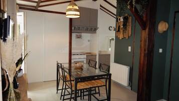 A dining area with a glass table and chairs, a hanging light fixture, and a kitchen in the background.