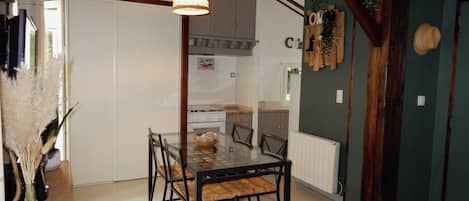 A dining area with a glass table and chairs, a hanging light fixture, and a kitchen in the background.