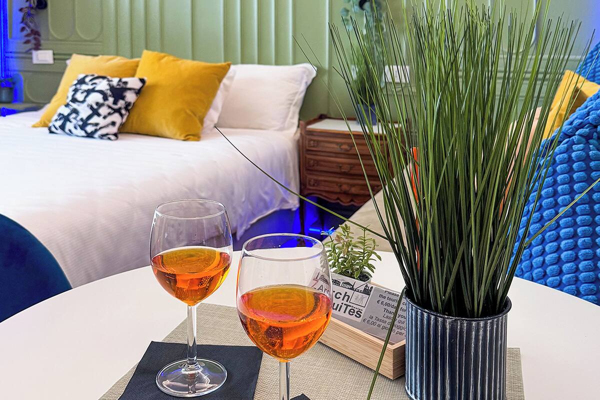 Una cama bien hecha con un almohadón blanco y amarillo, dos vasos de bebida color ámbar sobre una mesa, y una planta en maceta con hojas verdes largas.