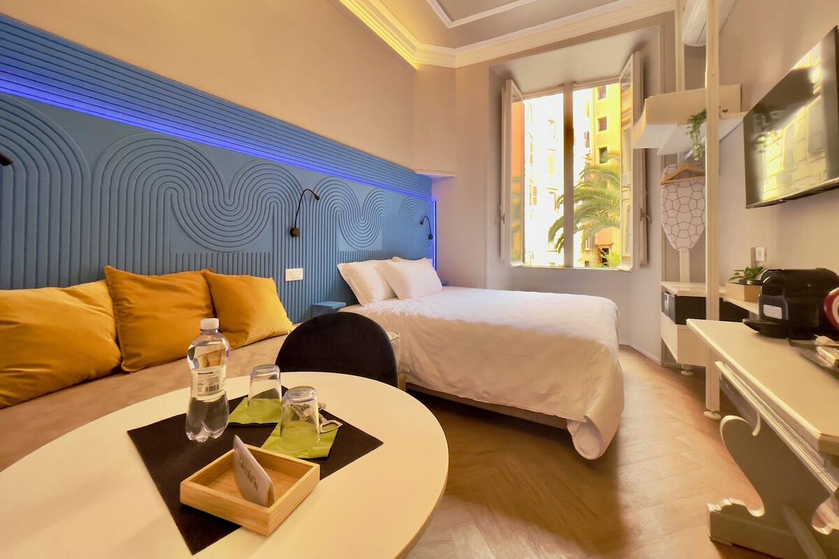 Habitación de hotel con cama, sofá, mesa con una botella y vasos, y ventana con vistas a edificios.