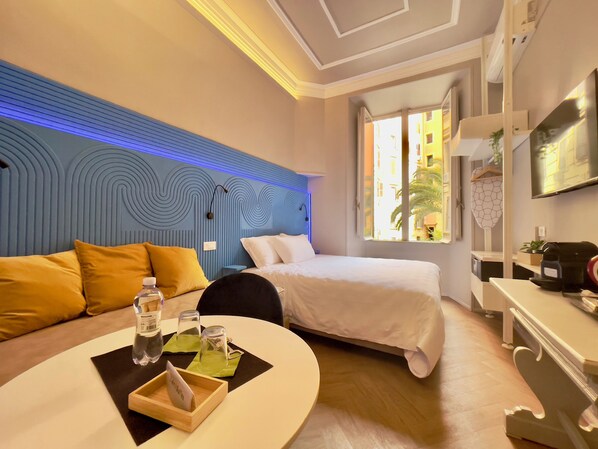 Habitación de hotel con cama, sofá, mesa con una botella y vasos, y ventana con vistas a edificios.