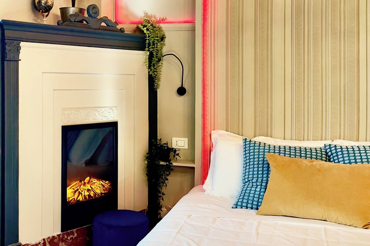 Un dormitorio con chimenea, una cama con almohadones estampados, un otoman azul y una planta.