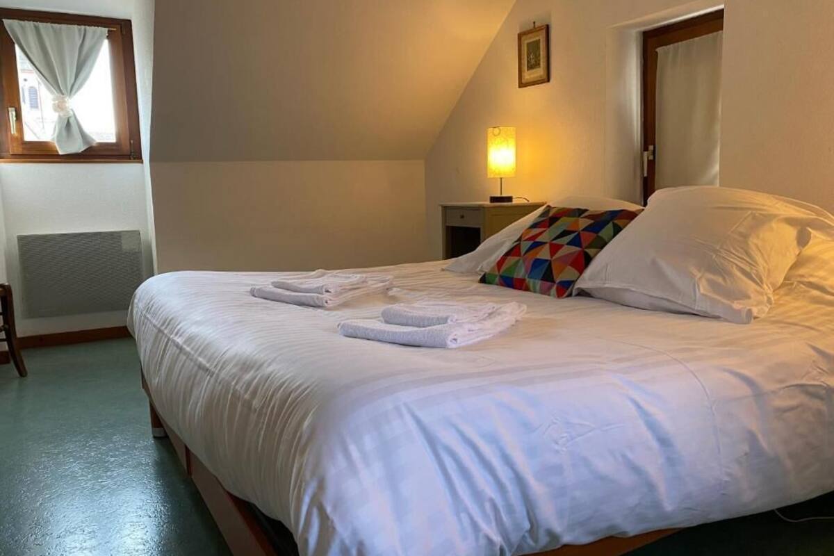 Un lit bien fait, avec du linge de lit et des serviettes blanches, un coussin coloré, une lampe de chevet et un tableau encadré au mur.