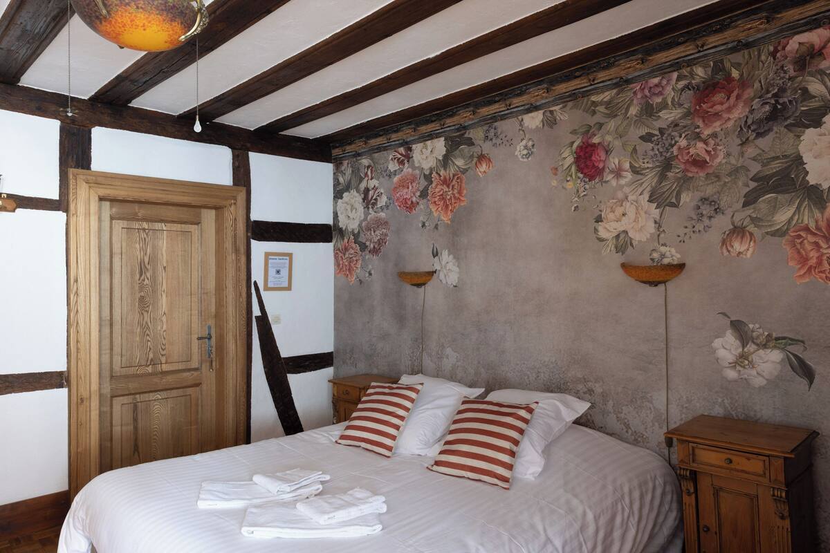 Une chambre à coucher avec un grand lit, des poutres en bois, un papier peint à motifs floraux et une armoire en bois.
