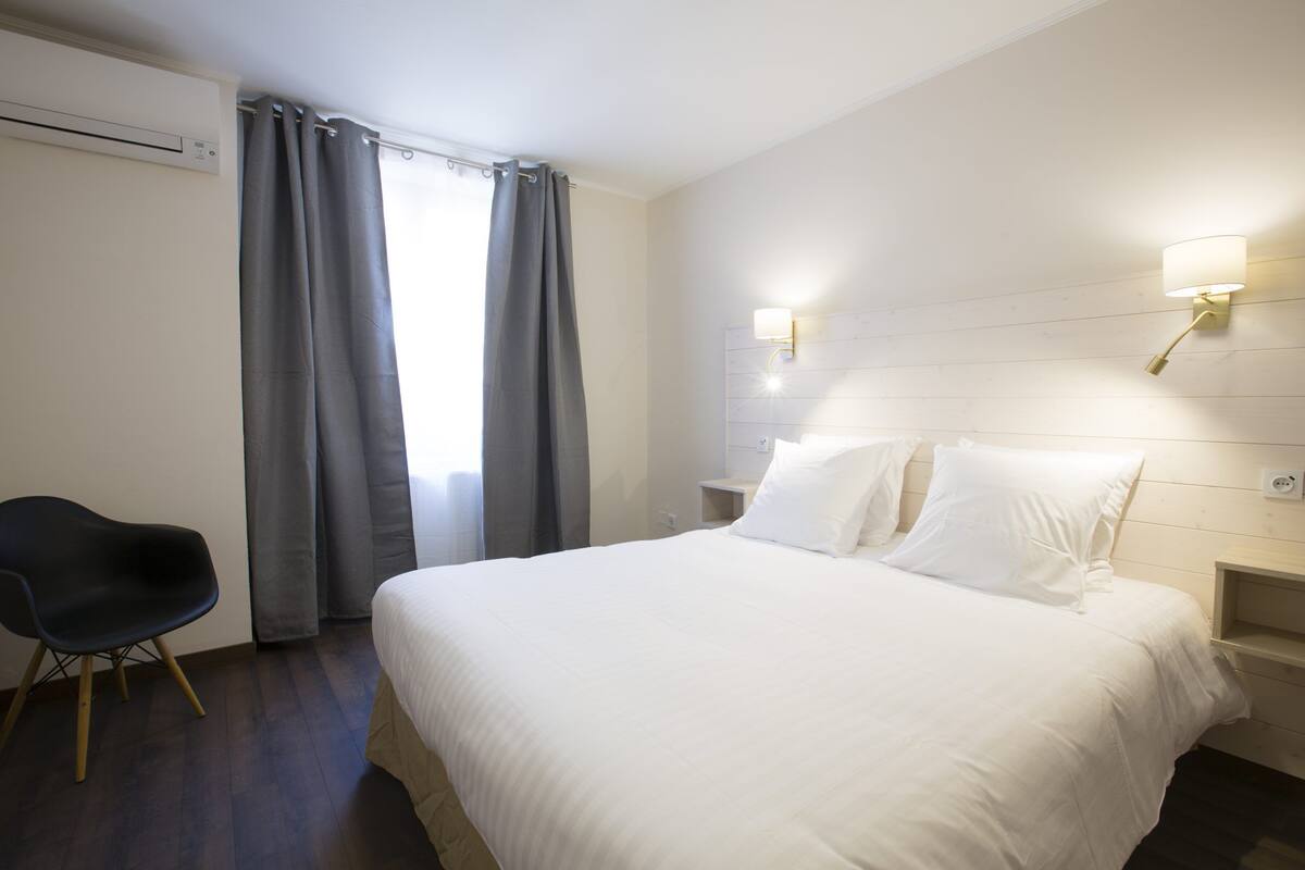 Une chambre d’hôtel avec un lit, une chaise, une table de chevet et un luminaire fixé au mur.