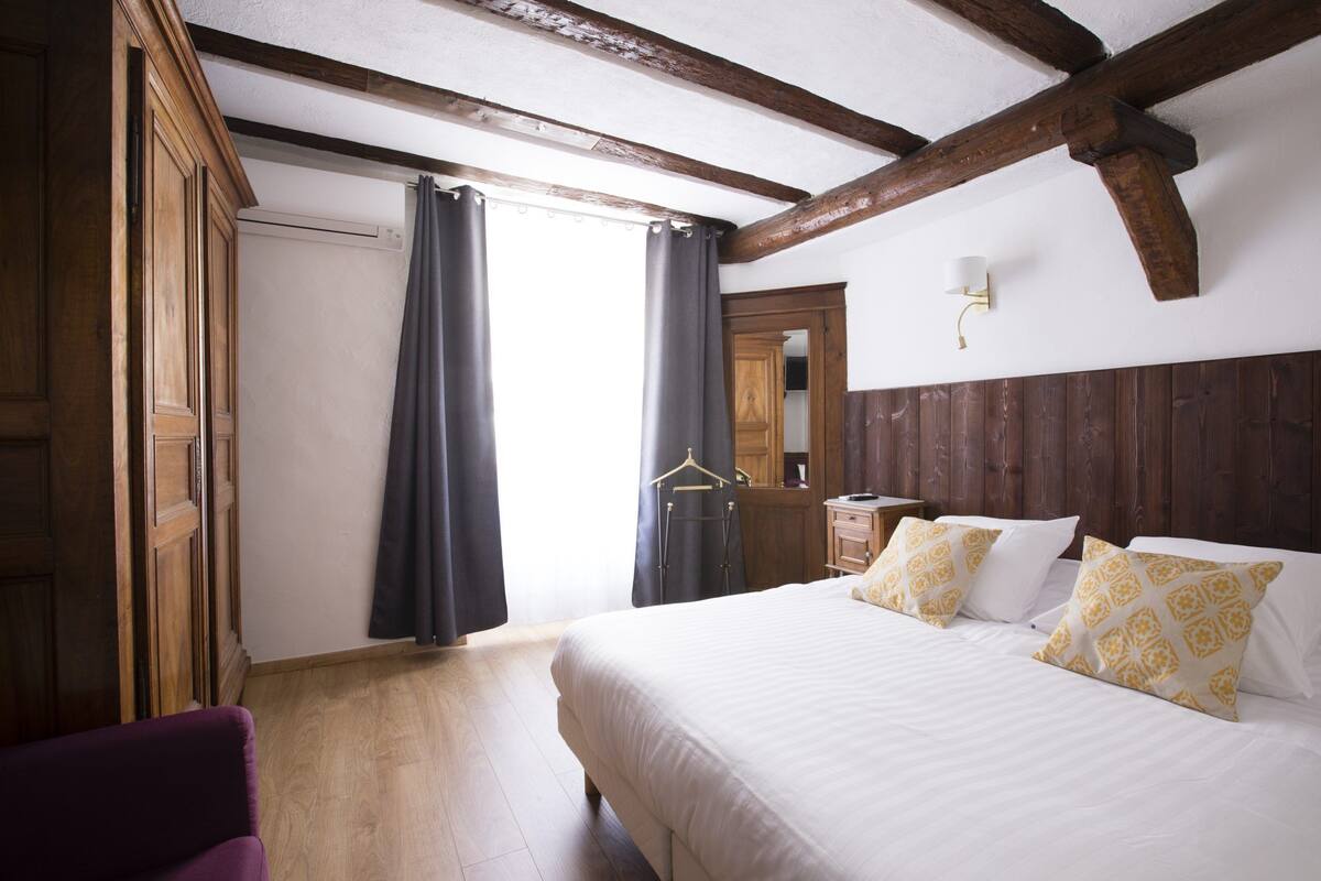 Une chambre avec un plafond en bois, un lit agrémenté d’oreillers blancs et à motifs, une armoire en bois et un porte-manteau suspendu.