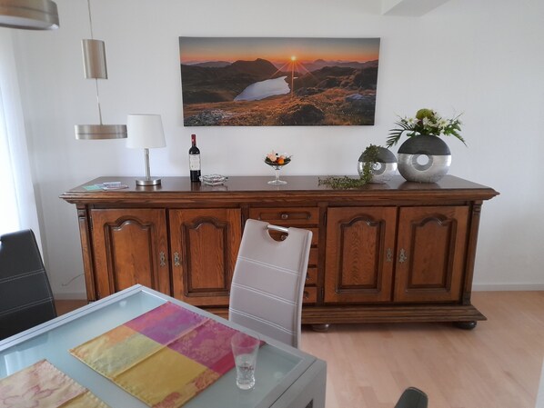 Uma sala de estar moderna com um armário de madeira, uma mesa de centro de vidro com jogos americanos coloridos, uma garrafa de vinho e uma planta decorativa.