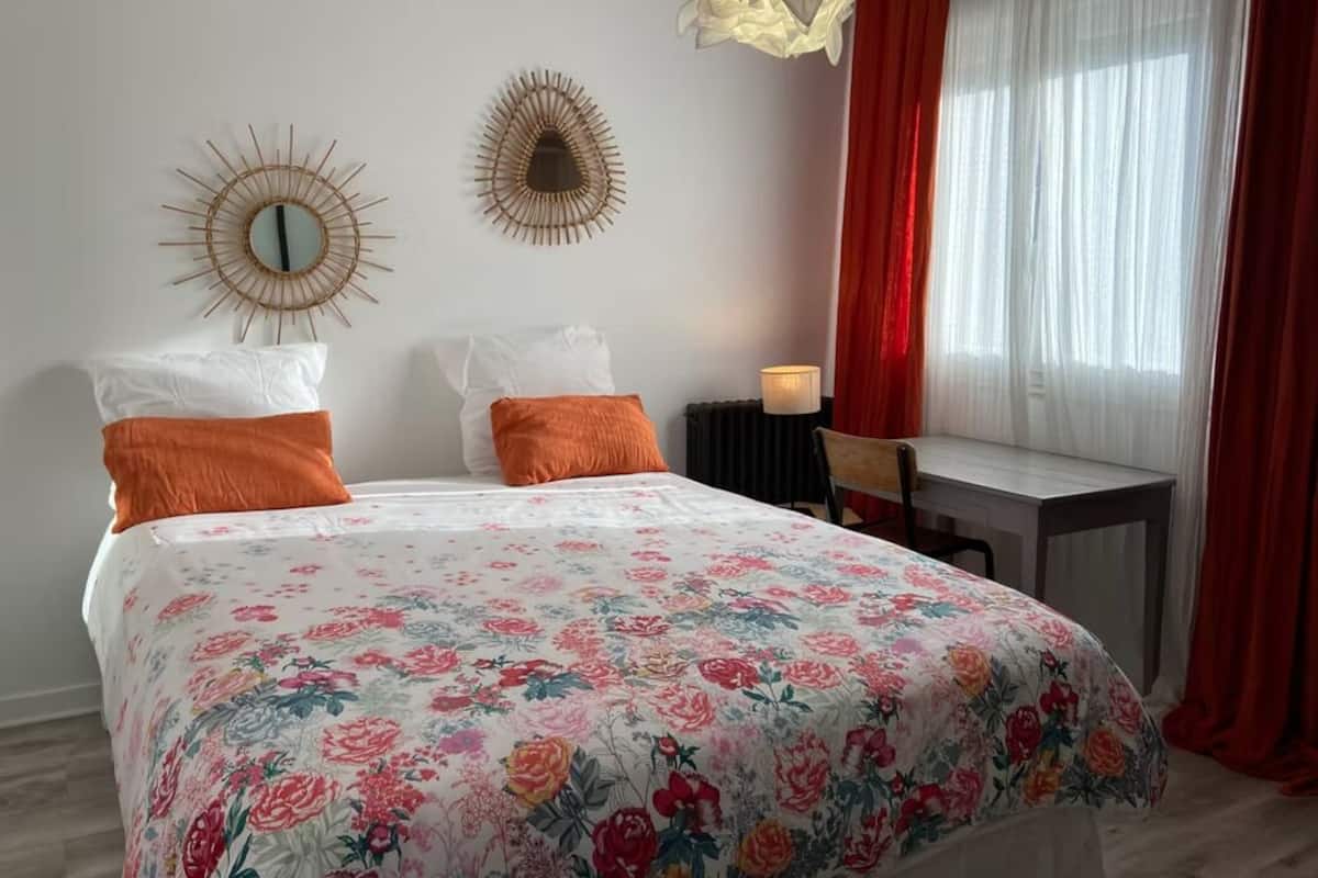Un dormitorio con colcha floral, dos espejos en la pared, un escritorio con silla y una ventana con cortinas rojas.