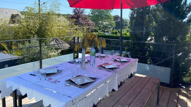 Un espace de restauration en plein air avec une table dressée pour quatre personnes, un parasol rouge et une vue sur la verdure et les bâtiments.