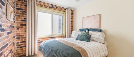 A bedroom with a brick wall, a large window with curtains, a bed with striped bedding, and a ceiling fan.