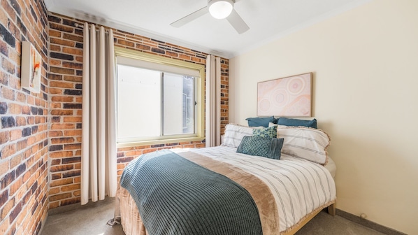 A bedroom with a brick wall, a large window with curtains, a bed with striped bedding, and a ceiling fan.