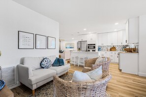 A modern living room with a sofa and wicker chairs, a kitchen with white cabinets, and framed pictures on the wall.