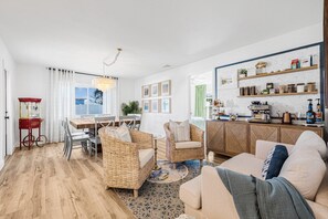 A spacious living room with a dining area, a kitchen with open shelving, and a view through a large window.