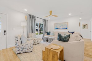 A modern living room with a beige sofa, patterned armchair, and ottoman, a wooden coffee table, and a ceiling fan.