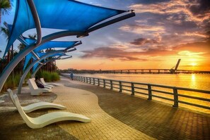 A waterfront promenade with modern sun loungers and blue canopies, a paved walkway, and a sunset view.