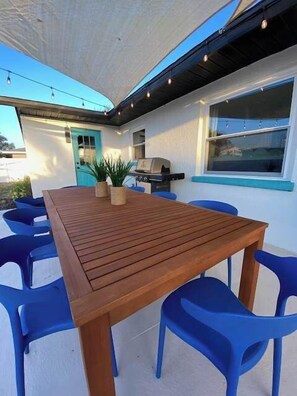 Um pátio externo com mesa de madeira e cadeiras de plástico azul, churrasqueira e um vaso com planta.
