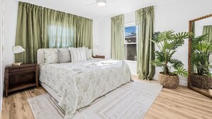 A bedroom with a bed, a nightstand, a bedside lamp, a ceiling fan, a window with curtains, a potted plant, and a mirror. - Castaway Cottage: Near IMG and Beaches (Bradenton)