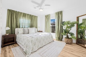 A bedroom with a bed, a nightstand, a bedside lamp, a ceiling fan, a window with curtains, a potted plant, and a mirror. - Castaway Cottage: Near IMG and Beaches (Bradenton)