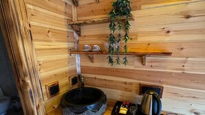 Interior de una cabaña de madera con un lavabo de piedra, una tetera y una repisa con tazas.