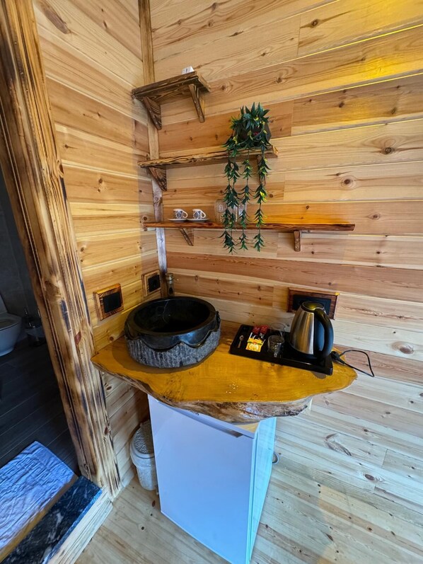 A wooden cabin interior with a stone basin, a kettle, and a shelf with mugs.