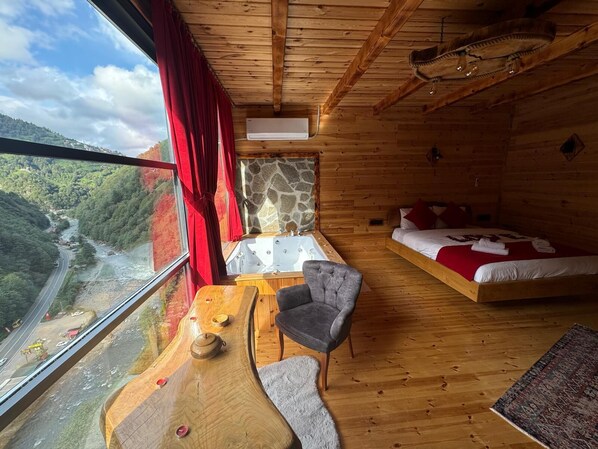 A wooden cabin with a bed, a sofa, a table, and a view of a river and mountains.