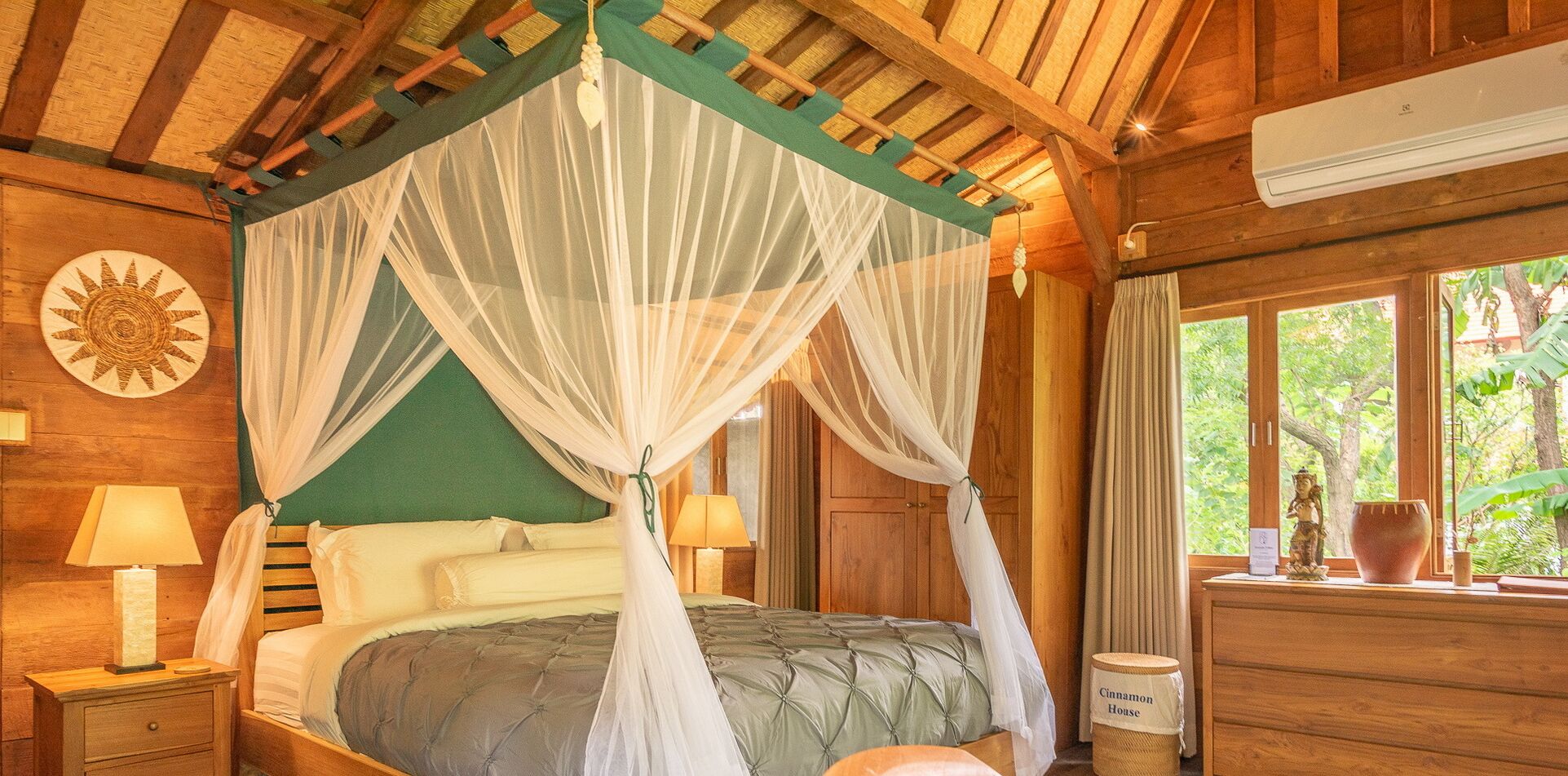 A room with a bed under a canopy, wooden walls, a nightstand with a lamp, a dresser, and a window with a view of greenery.