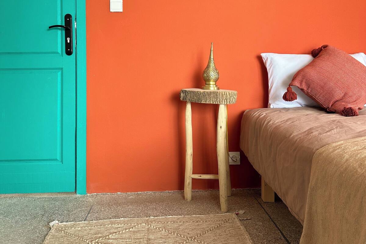 A room with a teal door, an orange wall, a small wooden table with a lamp, a bed with a beige bedspread and a pink pillow, and a beige rug.