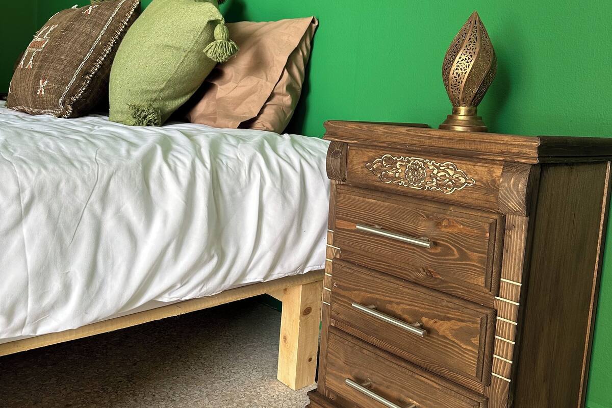 A bedroom with a bed against a green wall, a wooden dresser with drawers, and a patterned rug.