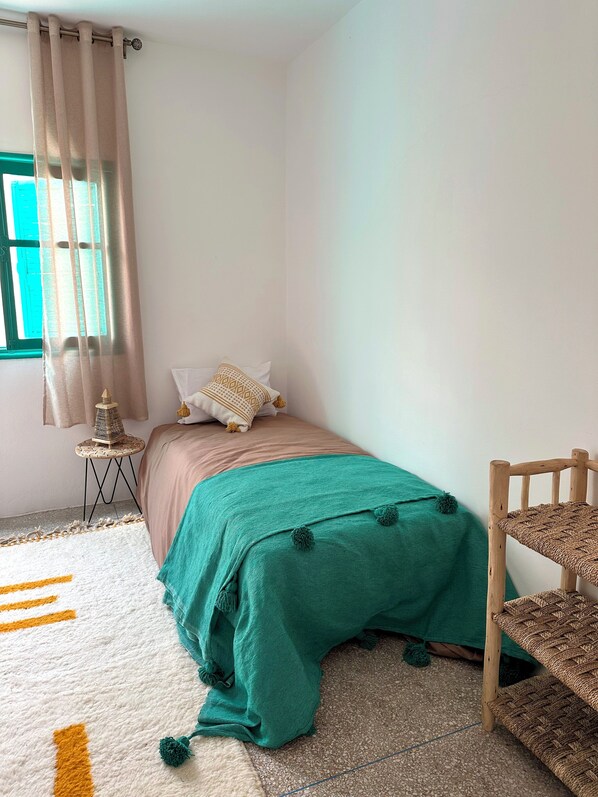 A bedroom with a bed, a small side table, a chair, and a window with curtains. - Dar Al-Andalus (Tamraght)