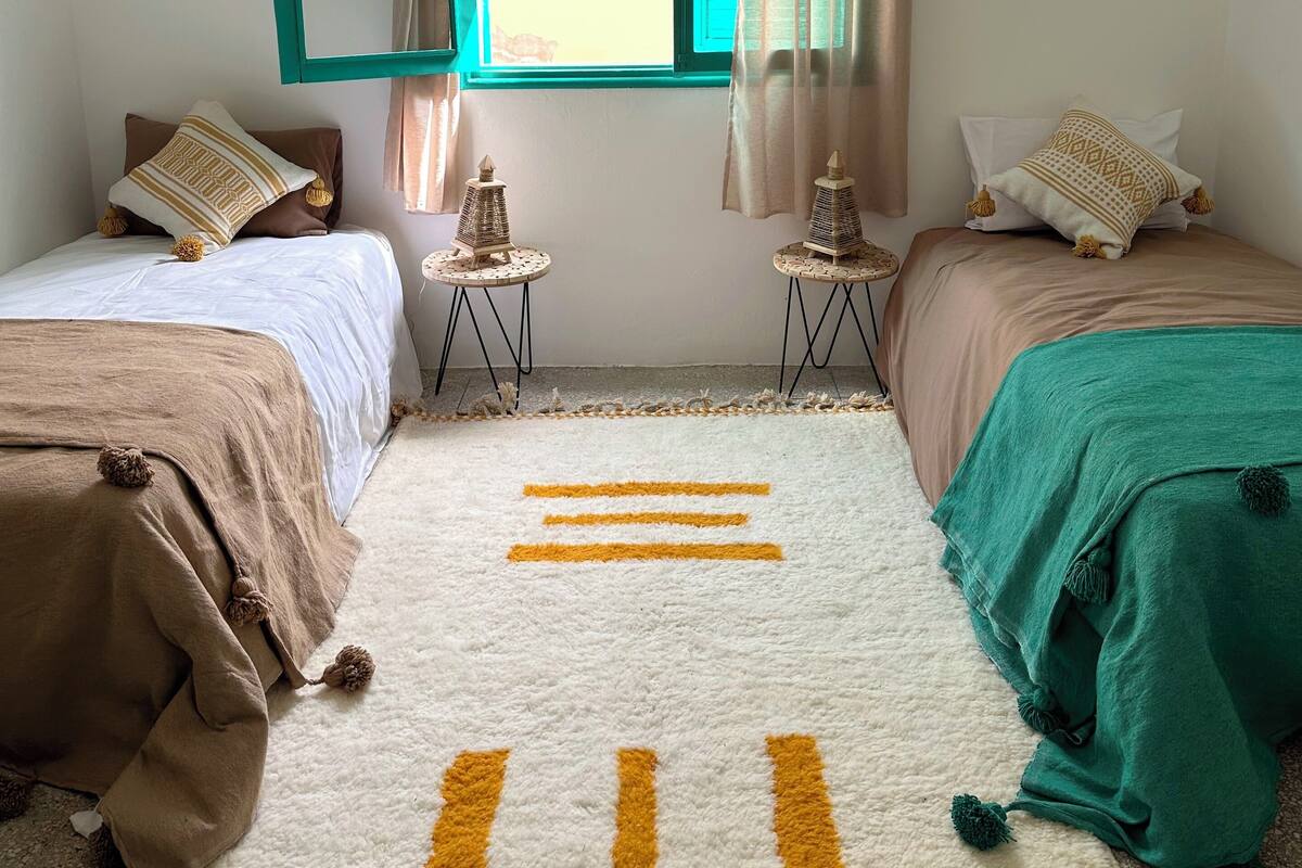 A room with two beds, a window with curtains, a small table with a lamp, and a patterned rug.