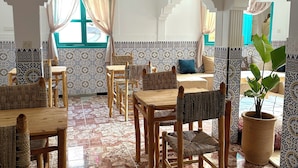 A dining area with wooden tables and chairs, patterned tile walls, and a large potted plant. - Dar Al-Andalus (Tamraght)