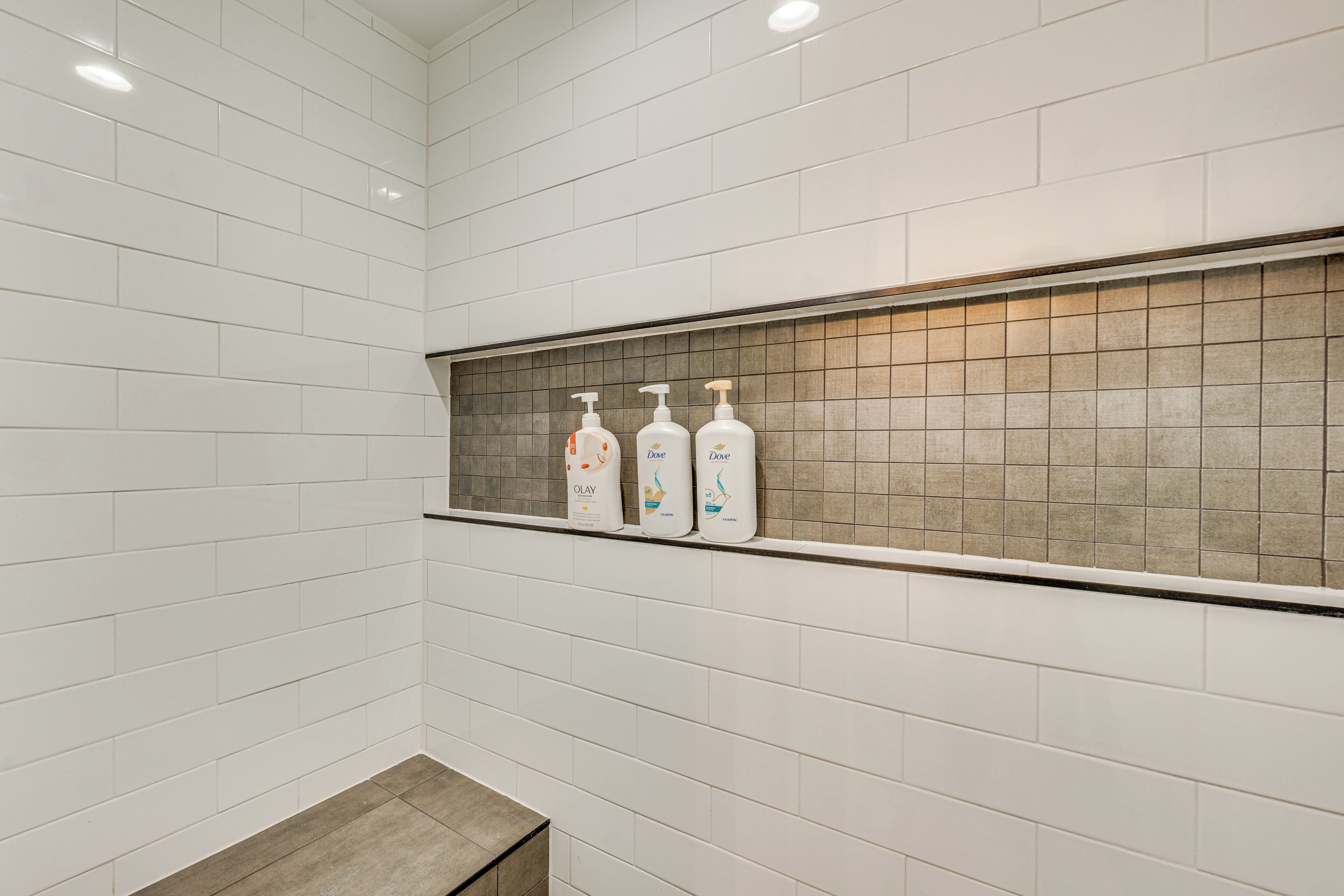 Une salle de bain avec des murs recouverts de carreaux blancs, une étagère sur laquelle reposent trois savons Dove, et un banc.