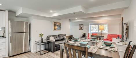 A modern kitchen with a stainless steel refrigerator, a dining area with a wooden table and chairs, a gray sofa, and a red couch.