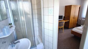 Ein Badezimmer mit Dusche, Waschbecken und Toilette. Im angrenzenden Raum befindet sich ein Holzschrank und ein Bett.