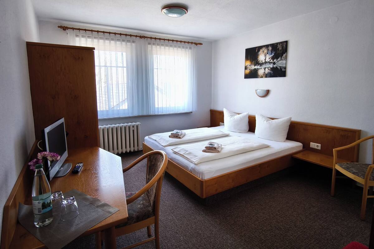 Ein Hotelzimmer mit einem Holzbett, zwei weißen Kissen, einem Nachttisch mit Flasche und Gläsern, einem Sessel, einem Schreibtisch mit Computer und einem Fenster mit Vorhängen.