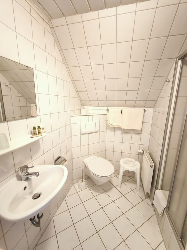 Un baño con baldosas blancas, un lavabo, un inodoro, un taburete pequeño y un espejo.
