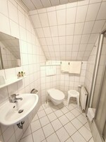 Um banheiro com azulejos brancos, pia, vaso sanitário, um pequeno banquinho e um espelho.